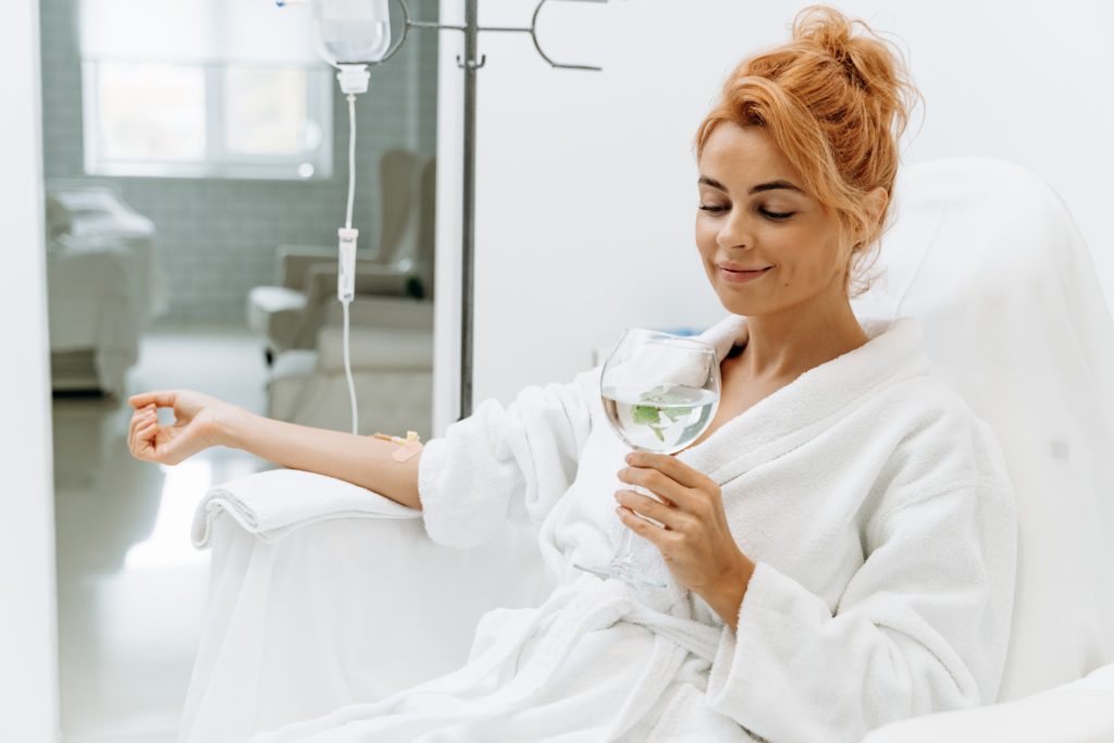 Close up of calm female patient sitting with tube and needle during IV infusion. Girl holding glass with lemon water and relaxing | Aviva Medical in Centennial, CO