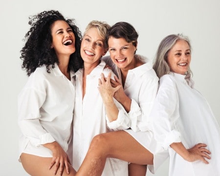 Four women different age smiling an looking happy and relaxed. Wearing white shirts against gray background. Emsella Chair, Aviva Med Spa, Centennial, CO